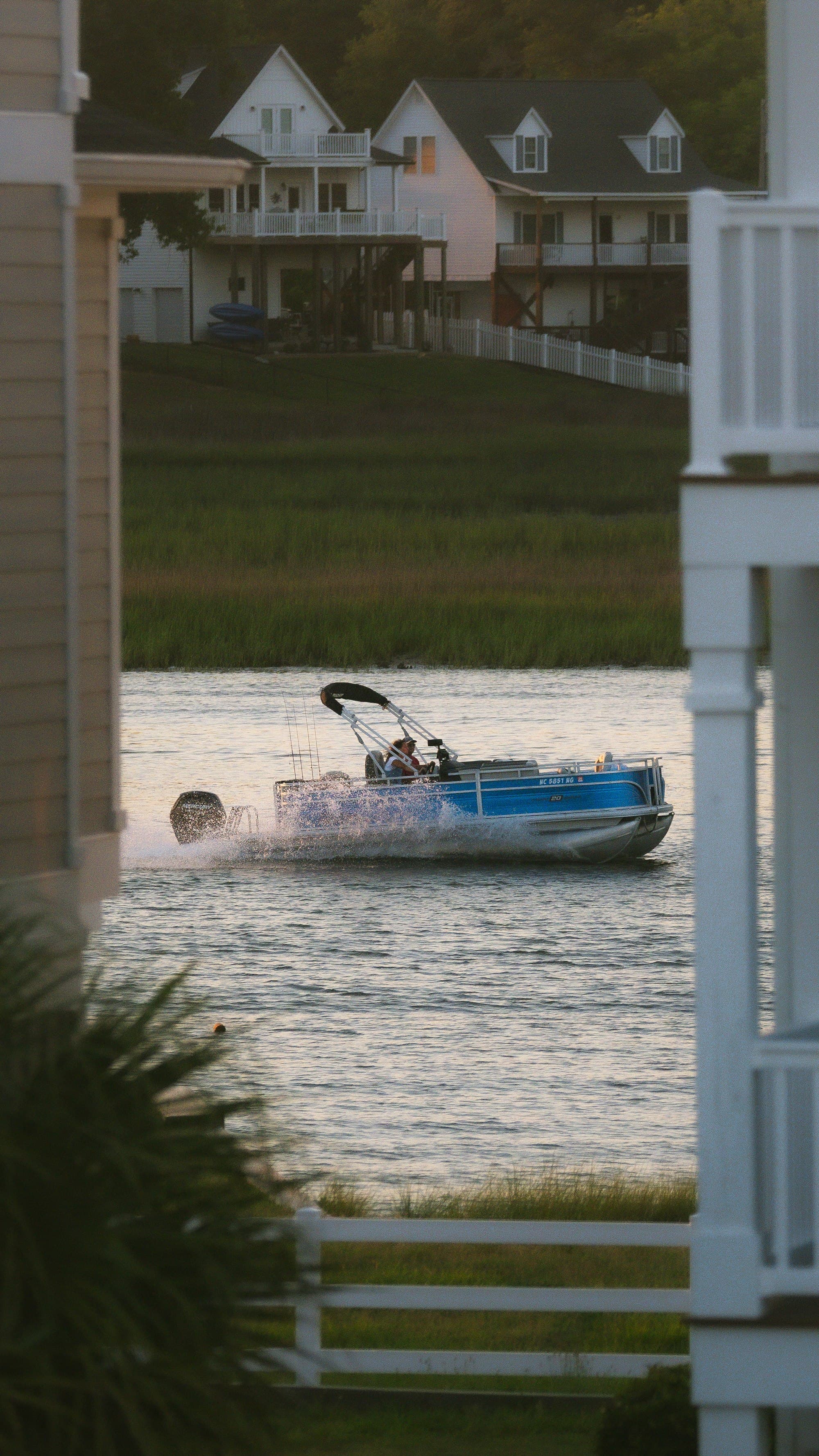 A blue fishing pontoon boat with rods in its holders running across a calm waterway